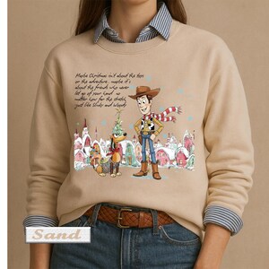 May include: A sand-colored crewneck sweatshirt featuring a graphic of Woody and Slinky Dog in a snowy village. The text reads: Maybe Christmas isn't about the toys or the adventure, maybe it's about the friends who never let go of your hand no matter how far the stretch, just like Slinky and Woody.