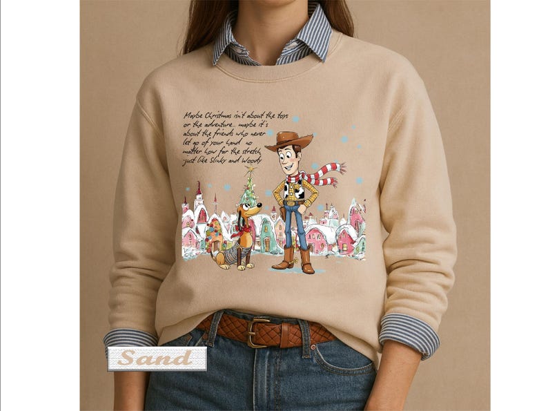May include: A sand-colored crewneck sweatshirt featuring a graphic of Woody and Slinky Dog in a snowy village. The text reads: Maybe Christmas isn't about the toys or the adventure, maybe it's about the friends who never let go of your hand no matter how far the stretch, just like Slinky and Woody.