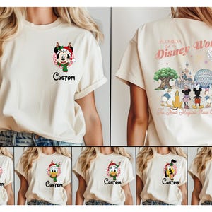 May include: A cream-coloured t-shirt featuring a small graphic on the front left chest of a Disney character in a holiday hat and the word Custom. The back displays a large graphic of Disney World landmarks and characters with the text Florida Est 1971.