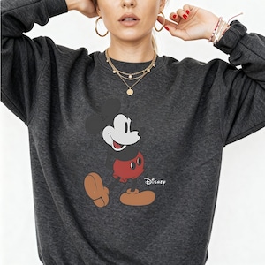 May include: A dark charcoal gray crewneck sweatshirt featuring a graphic print of Mickey Mouse on the front. The character is depicted with a black head, red shorts, and tan shoes. The word Disney is printed in white script next to the character.