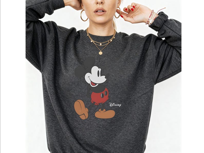May include: A dark charcoal gray crewneck sweatshirt featuring a graphic print of Mickey Mouse on the front. The character is depicted with a black head, red shorts, and tan shoes. The word Disney is printed in white script next to the character.