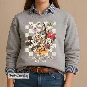 May include: A heather sport gray crewneck sweatshirt featuring a graphic of Mickey Mouse, Minnie Mouse, Donald Duck, Daisy Duck, Goofy, and Pluto over a cream and white checkered background. The text reads MICKEY & CO. EST. 1928 in a brown font.