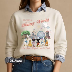 May include: A cream-colored crewneck sweatshirt featuring a graphic print of Disney characters and landmarks. The text reads Florida Est. 1971 Disney World and The Most Magical Place On Earth. The design includes Mickey, Minnie, Donald, Daisy, Pluto, and Goofy.