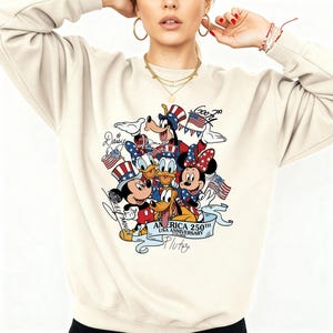 May include: A cream-colored crewneck sweatshirt featuring a graphic print of Disney characters including Mickey Mouse, Minnie Mouse, Donald Duck, Daisy Duck, Goofy, and Pluto wearing patriotic hats, with the text America 250th USA Anniversary.