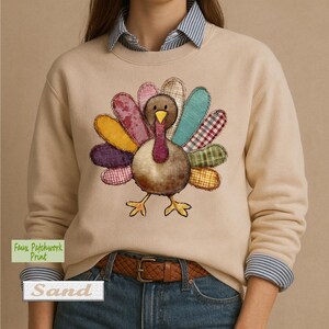 May include: A beige crewneck sweatshirt featuring a central graphic of a turkey with colorful faux patchwork feathers in shades of yellow, pink, teal, red, and green. The turkey has a brown body, a yellow beak, and a red wattle.