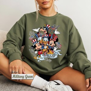 May include: A military green crewneck sweatshirt featuring a graphic print of Disney characters including Mickey Mouse, Minnie Mouse, Donald Duck, Goofy, and Pluto wearing patriotic hats. The text reads America 250th USA Anniversary 1776-2026.