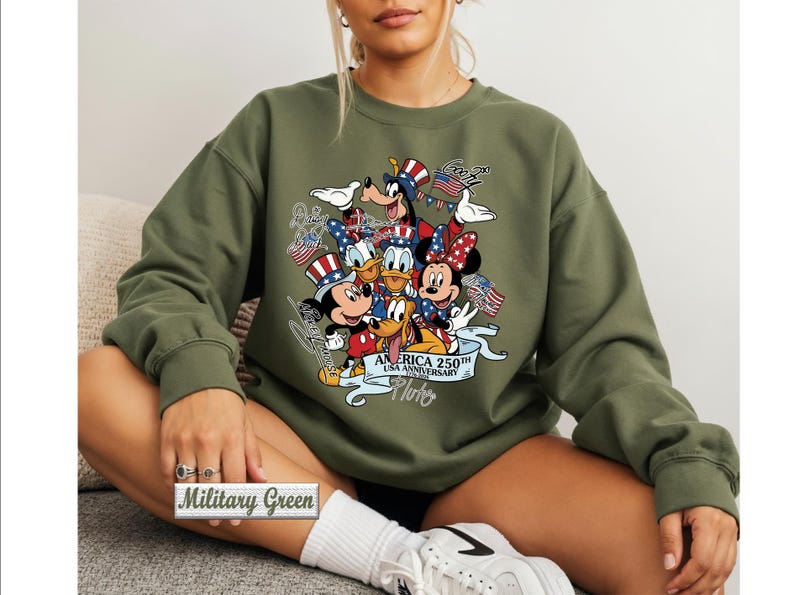 May include: A military green crewneck sweatshirt featuring a graphic print of Disney characters including Mickey Mouse, Minnie Mouse, Donald Duck, Goofy, and Pluto wearing patriotic hats. The text reads America 250th USA Anniversary 1776-2026.