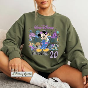 Op de afbeelding: Een olijfgroene sweater met ronde hals en een grafische print van Mickey Mouse in een blauw pak, omringd door kleurrijke illustraties van Disneyland-bezienswaardigheden, met de tekst Walt Disney, Disneyland en het getal 20 in paars en zwart.