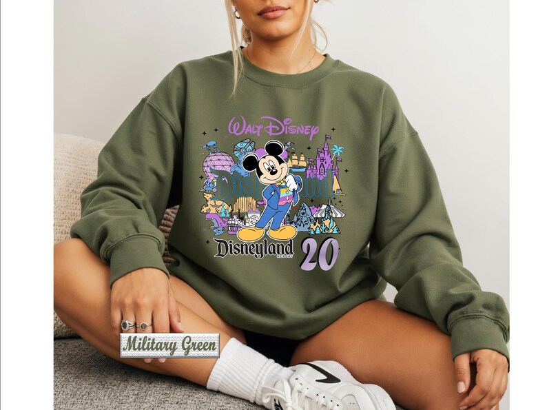 Op de afbeelding: Een olijfgroene sweater met ronde hals en een grafische print van Mickey Mouse in een blauw pak, omringd door kleurrijke illustraties van Disneyland-bezienswaardigheden, met de tekst Walt Disney, Disneyland en het getal 20 in paars en zwart.