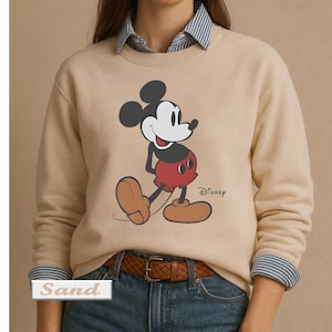 May include: A sand-colored crewneck sweatshirt featuring a graphic of Mickey Mouse in red shorts and brown shoes. The Disney logo is printed in black next to the character. The sweatshirt is worn over a blue and white striped collared shirt.