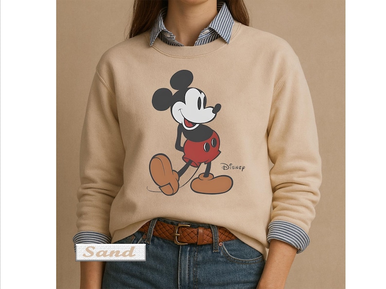May include: A sand-colored crewneck sweatshirt featuring a graphic of Mickey Mouse in red shorts and brown shoes. The Disney logo is printed in black next to the character. The sweatshirt is worn over a blue and white striped collared shirt.