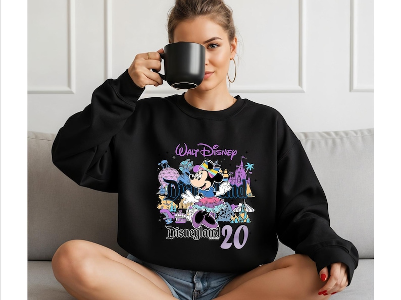 Op de afbeelding: Een zwart sweatshirt met ronde hals en een kleurrijke grafische print van Minnie Mouse, de tekst Walt Disney Disneyland en het getal 20. Het ontwerp bevat illustraties van diverse parkmonumenten in paarse, blauwe en gele tinten.
