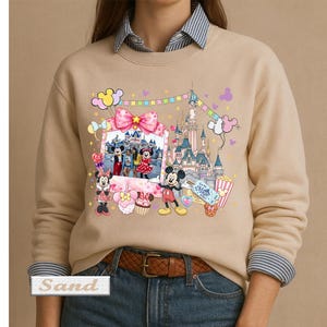 May include: A sand-colored crewneck sweatshirt featuring a graphic print of a castle, Mickey Mouse, and Minnie Mouse. The design includes a photo frame, colorful balloons, a popcorn bucket, and sweets. The word Sand is printed in a small box.