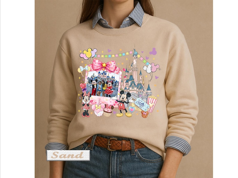 May include: A sand-colored crewneck sweatshirt featuring a graphic print of a castle, Mickey Mouse, and Minnie Mouse. The design includes a photo frame, colorful balloons, a popcorn bucket, and sweets. The word Sand is printed in a small box.