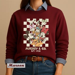 May include: A maroon crewneck sweatshirt featuring a graphic print of Mickey Mouse, Minnie Mouse, Donald Duck, Daisy Duck, Goofy, and Pluto over a white and maroon checkerboard pattern. The text MICKEY & CO. EST. 1928 is printed below the characters.