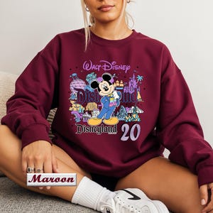 Op de afbeelding: Een bordeauxrood sweatshirt met lange mouwen en ronde hals, voorzien van een grafische print van Mickey Mouse in een blauw pak, de tekst Walt Disney Disneyland en het getal 20. Het ontwerp bevat kleurrijke illustraties van attracties.