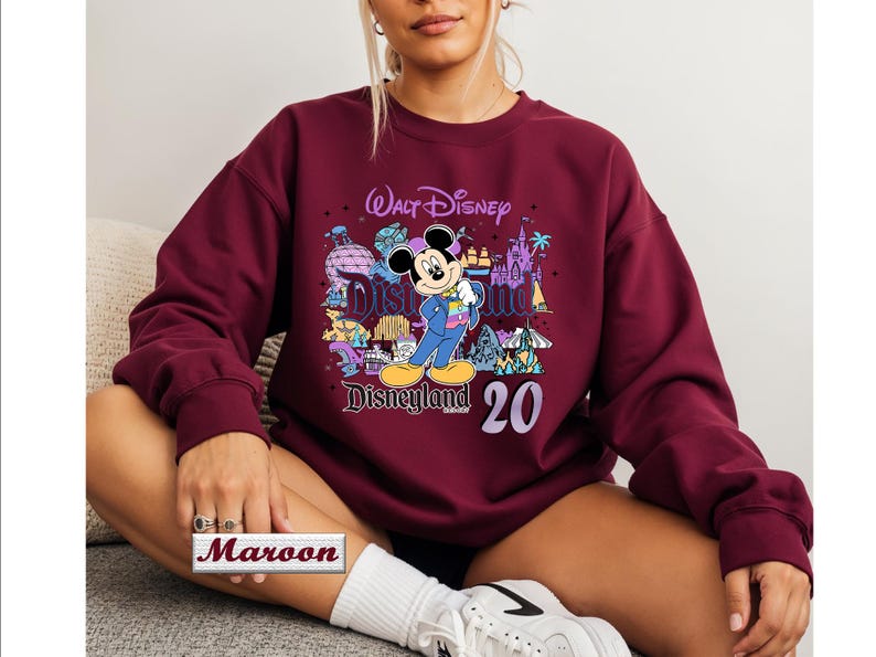 Op de afbeelding: Een bordeauxrood sweatshirt met lange mouwen en ronde hals, voorzien van een grafische print van Mickey Mouse in een blauw pak, de tekst Walt Disney Disneyland en het getal 20. Het ontwerp bevat kleurrijke illustraties van attracties.
