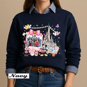 May include: A navy blue crewneck sweatshirt featuring a colorful graphic print of a castle, Mickey Mouse, Minnie Mouse, a photo frame with characters, balloons, a cupcake, popcorn, and a ticket with the text Let the Magic Begin printed on the front.