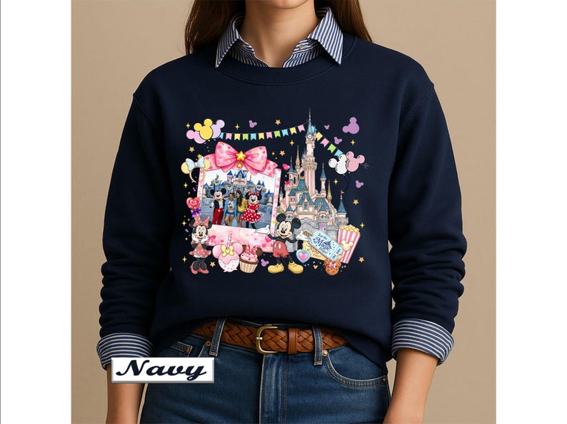 May include: A navy blue crewneck sweatshirt featuring a colorful graphic print of a castle, Mickey Mouse, Minnie Mouse, a photo frame with characters, balloons, a cupcake, popcorn, and a ticket with the text Let the Magic Begin printed on the front.
