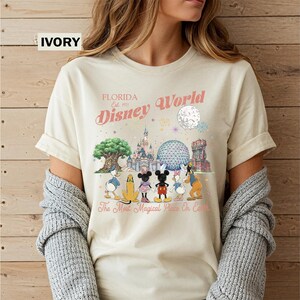 May include: An ivory t-shirt featuring a graphic print of Disney World landmarks and characters including Mickey Mouse, Minnie Mouse, Donald Duck, Daisy Duck, Pluto, and Goofy. The text reads Florida Est. 1971 Disney World and The Most Magical Place On Earth.