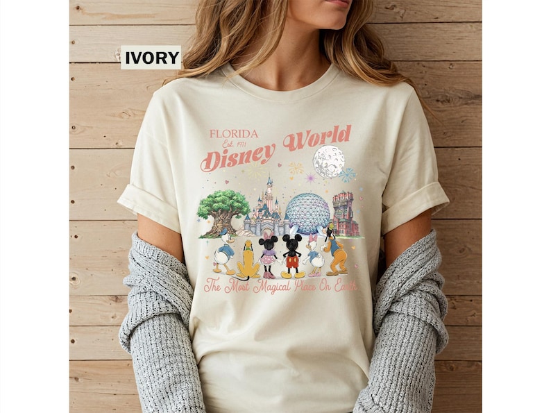 May include: An ivory t-shirt featuring a graphic print of Disney World landmarks and characters including Mickey Mouse, Minnie Mouse, Donald Duck, Daisy Duck, Pluto, and Goofy. The text reads Florida Est. 1971 Disney World and The Most Magical Place On Earth.