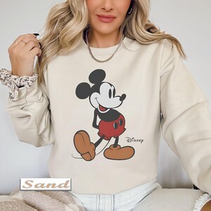 May include: A sand-colored crewneck sweatshirt featuring a graphic print of Mickey Mouse in his classic black, white, and red color scheme. The character is depicted standing with a smile, and the word Disney is printed in black near the feet.