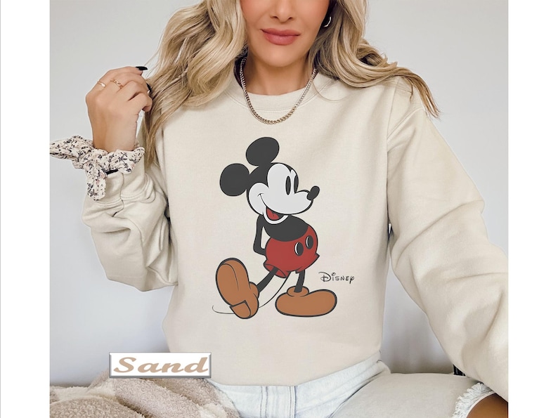 May include: A sand-colored crewneck sweatshirt featuring a graphic print of Mickey Mouse in his classic black, white, and red color scheme. The character is depicted standing with a smile, and the word Disney is printed in black near the feet.
