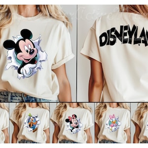 May include: A cream-colored short-sleeve t-shirt featuring a graphic of Mickey Mouse emerging from a torn paper effect on the front. The back of the shirt displays the word DISNEYLAND in bold black lettering. Other versions show characters.