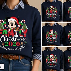 May include: A navy blue crewneck sweatshirt featuring a graphic print of Mickey Mouse in a Santa hat, surrounded by Christmas trees, candy canes, and the text "FAMILY Christmas 2026 making memories together" in red, white, and green colors.