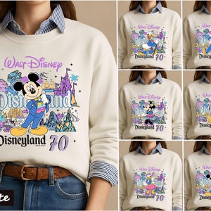 Op de afbeelding: Een cr&egrave;mekleurig sweatshirt met ronde hals en een grafische print van Walt Disney Disneyland 70 met diverse Disney-figuren zoals Mickey Mouse, Donald Duck en Goofy, gedragen over een blauw-wit gestreept overhemd.