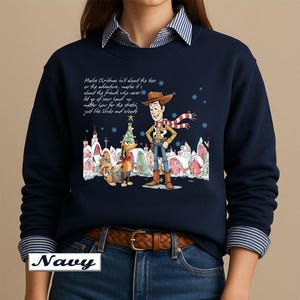 May include: A navy blue long-sleeve sweatshirt featuring a graphic of Woody and Slinky Dog from Toy Story in a snowy village setting. The text reads: Maybe Christmas isn't about the toys or the adventure, maybe it's about the friends who never let go of your hand no matter how far the stretch, just like Slinky and Woody.