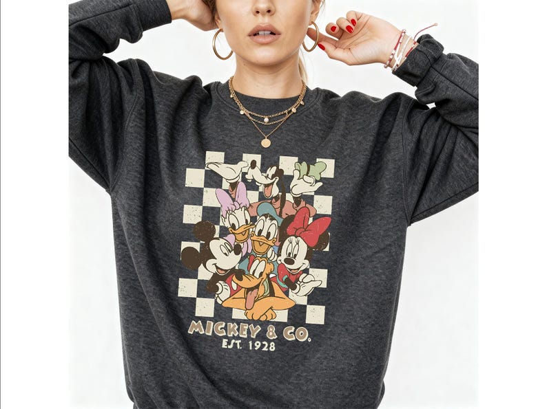 May include: A dark charcoal gray long-sleeve sweatshirt featuring a graphic print of Mickey Mouse, Minnie Mouse, Donald Duck, Daisy Duck, Goofy, and Pluto against a cream and gray checkered background with the text Mickey & Co. Est. 1928.