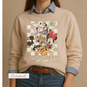 May include: A sand-colored crewneck sweatshirt featuring a graphic print of Mickey Mouse, Minnie Mouse, Donald Duck, Daisy Duck, Goofy, and Pluto over a white checkered background. The text MICKEY & CO. EST. 1928 is printed below the characters.