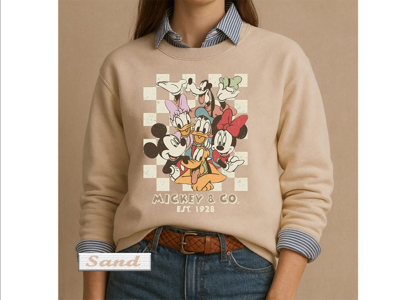 May include: A sand-colored crewneck sweatshirt featuring a graphic print of Mickey Mouse, Minnie Mouse, Donald Duck, Daisy Duck, Goofy, and Pluto over a white checkered background. The text MICKEY & CO. EST. 1928 is printed below the characters.