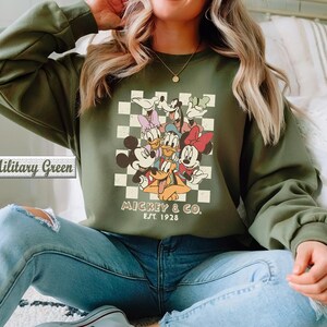 May include: A military green long-sleeve sweatshirt featuring a graphic print of Mickey Mouse, Minnie Mouse, Donald Duck, Daisy Duck, Goofy, and Pluto over a cream and green checkered background with the text Mickey & Co. Est. 1928.