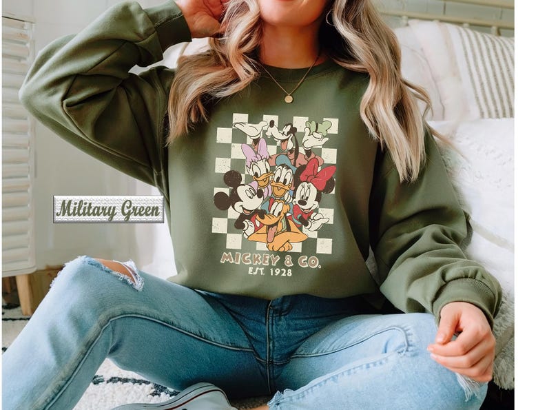 May include: A military green long-sleeve sweatshirt featuring a graphic print of Mickey Mouse, Minnie Mouse, Donald Duck, Daisy Duck, Goofy, and Pluto over a cream and green checkered background with the text Mickey & Co. Est. 1928.