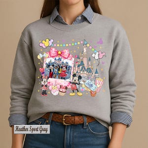 May include: A heather sport gray crewneck sweatshirt featuring a graphic print of Mickey and Minnie Mouse, a castle, balloons, and a framed photo. The design includes colorful bunting, a pink bow, and treats. The text "Heather Sport Gray" is visible.