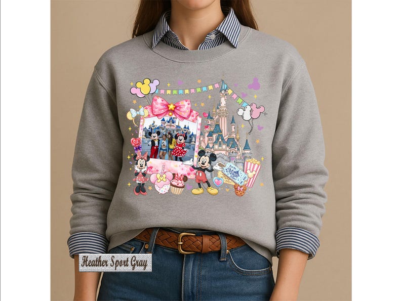 May include: A heather sport gray crewneck sweatshirt featuring a graphic print of Mickey and Minnie Mouse, a castle, balloons, and a framed photo. The design includes colorful bunting, a pink bow, and treats. The text "Heather Sport Gray" is visible.