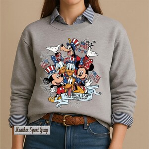 May include: A heather sport gray crewneck sweatshirt featuring a graphic of Disney characters including Mickey Mouse, Minnie Mouse, Donald Duck, Goofy, and Pluto wearing patriotic hats. The text reads America 250th USA Anniversary 1776-2026.