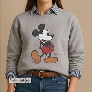 May include: A heather sport gray crewneck sweatshirt featuring a graphic of Mickey Mouse in his classic red shorts and tan shoes. The Disney logo is printed in black next to the character. The sweatshirt is worn over a blue and white striped shirt.