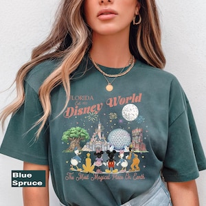May include: A blue spruce t-shirt featuring a graphic print of Disney World with the text Florida Est. 1971, Disney World, and The Most Magical Place On Earth. The design includes illustrations of the castle, park icons, and cartoon characters.