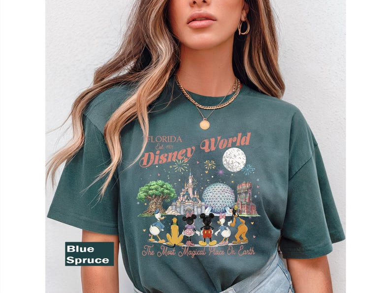 May include: A blue spruce t-shirt featuring a graphic print of Disney World with the text Florida Est. 1971, Disney World, and The Most Magical Place On Earth. The design includes illustrations of the castle, park icons, and cartoon characters.