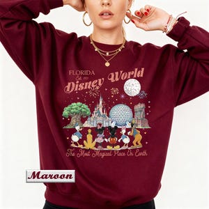 May include: A maroon crewneck sweatshirt featuring a graphic print of Disney World landmarks and characters. The text reads Florida Est. 1971 Disney World and The Most Magical Place On Earth. A small rectangular box in the corner contains the word Maroon.