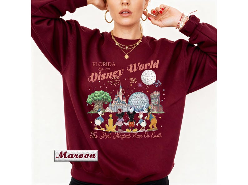 May include: A maroon crewneck sweatshirt featuring a graphic print of Disney World landmarks and characters. The text reads Florida Est. 1971 Disney World and The Most Magical Place On Earth. A small rectangular box in the corner contains the word Maroon.