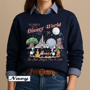 May include: A navy blue long-sleeve sweatshirt featuring a graphic print of Disney World landmarks and characters. The text reads Florida Est. 1971 Disney World and The Most Magical Place On Earth. The design includes a moon, fireworks, and icons.