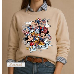 May include: A sand-colored crewneck sweatshirt featuring a graphic print of Disney characters including Mickey Mouse, Minnie Mouse, Donald Duck, Goofy, and Pluto wearing patriotic hats. The text reads America 250th USA Anniversary 1776-2026.