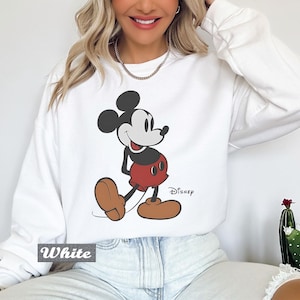 May include: A white long-sleeve crewneck sweatshirt featuring a graphic print of Mickey Mouse in his classic red shorts and brown shoes. The character is positioned centrally on the chest, with the word Disney printed in black script near the bottom right.