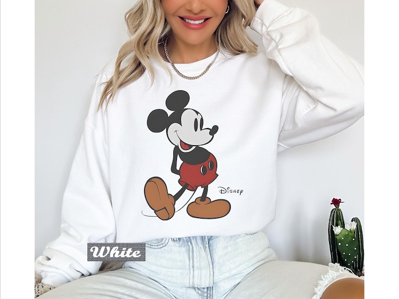 May include: A white long-sleeve crewneck sweatshirt featuring a graphic print of Mickey Mouse in his classic red shorts and brown shoes. The character is positioned centrally on the chest, with the word Disney printed in black script near the bottom right.