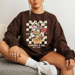 May include: A dark brown crewneck sweatshirt featuring a graphic print of Mickey Mouse, Minnie Mouse, Donald Duck, Daisy Duck, Goofy, and Pluto over a cream and brown checkerboard pattern with the text Mickey & Co. Est. 1928 printed below.