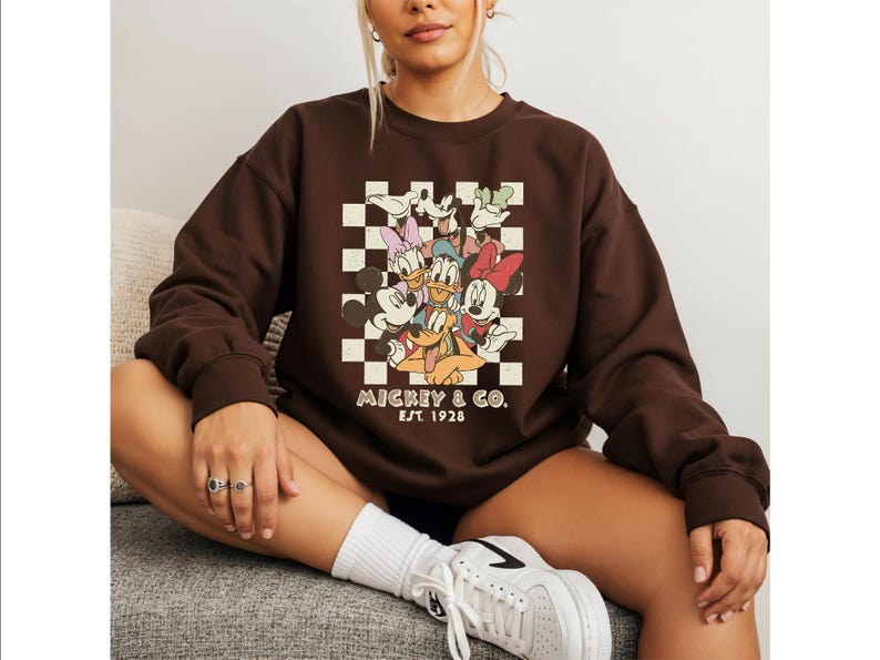 May include: A dark brown crewneck sweatshirt featuring a graphic print of Mickey Mouse, Minnie Mouse, Donald Duck, Daisy Duck, Goofy, and Pluto over a cream and brown checkerboard pattern with the text Mickey & Co. Est. 1928 printed below.